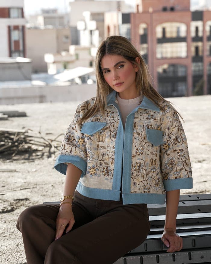 A cropped cream jacket with a floral pattern and light denim trim, worn seated on a rooftop